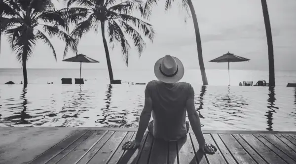 Person in a hat relaxing by a tropical resort pool with palm trees, for B2 First (FCE) speaking test imagery.